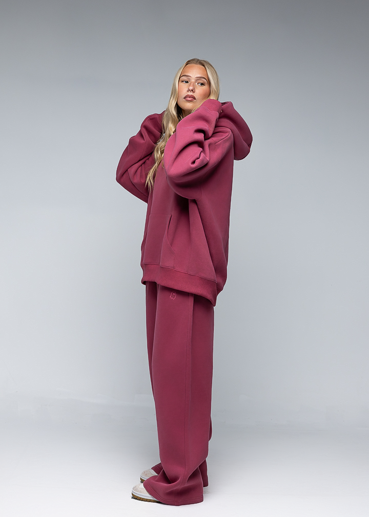 A woman modeling a matching burgundy lounge set consisting of an oversized hoodie and wide-leg sweatpants, photographed against a neutral gray studio background.
