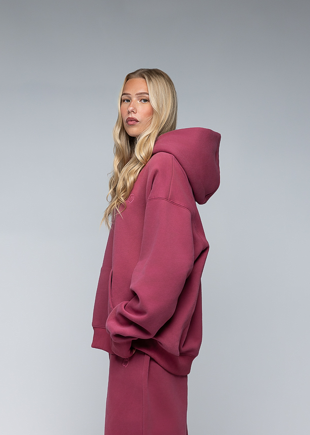 A blonde model wearing a matching dusty rose pink hooded sweatshirt and long skirt, shown in profile against a neutral gray studio background.