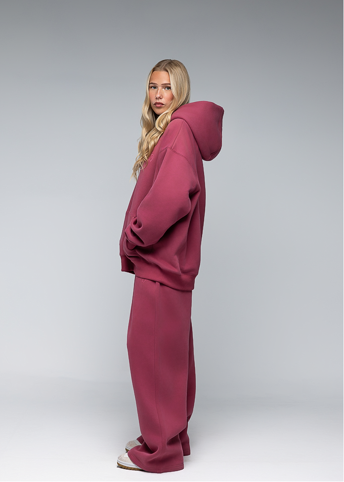 A woman modeling a matching dusty rose pink loungewear set consisting of an oversized hoodie and wide-leg sweatpants, photographed against a neutral gray studio background.