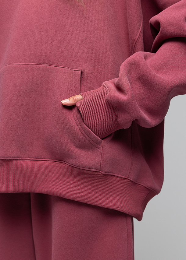 A close-up of a person wearing a matching dusty rose pink sweatshirt and sweatpants set, showing the front pocket of the sweatshirt and a hand resting inside the pocket.