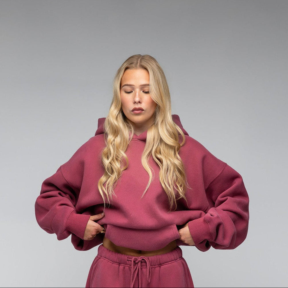 A blonde model wearing a matching dusty rose pink lounge set consisting of a cropped hoodie and wide-leg sweatpants, standing against a neutral gray studio background.