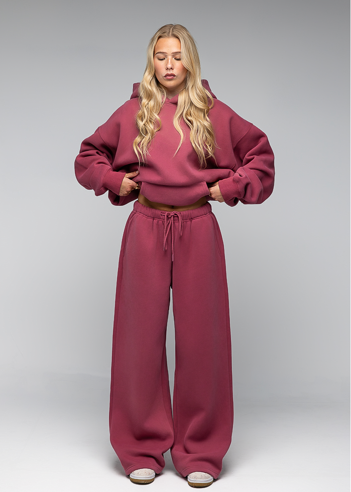 A blonde model wearing a matching dusty rose two-piece loungewear set, consisting of a cropped pullover hoodie and wide-leg sweatpants, standing against a neutral gray studio background.