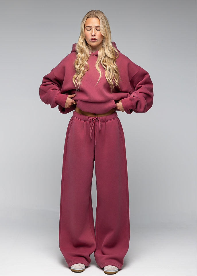 A blonde model wearing a matching dusty rose two-piece loungewear set, consisting of a cropped pullover hoodie and wide-leg sweatpants, standing against a neutral gray studio background.