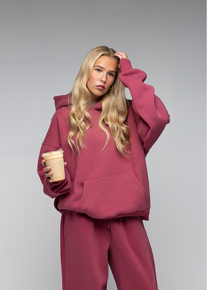 A blonde woman posing in a matching dusty rose pink lounge set, featuring an oversized pullover hoodie and matching sweatpants, holding a paper coffee cup against a neutral gray studio background.