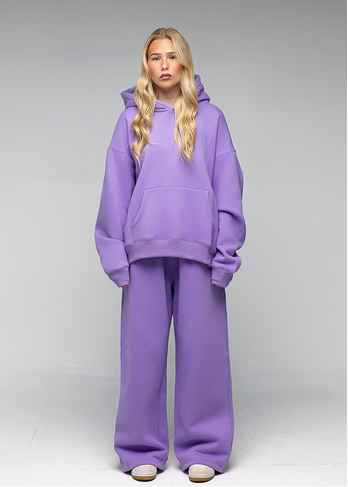 A blonde model wearing a matching lavender purple sweatshirt and wide-leg sweatpants set, standing against a neutral gray studio background.