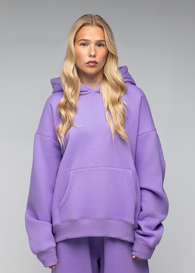 A blonde woman modeling an oversized lavender purple hoodie and matching sweatpants against a neutral gray studio background.
