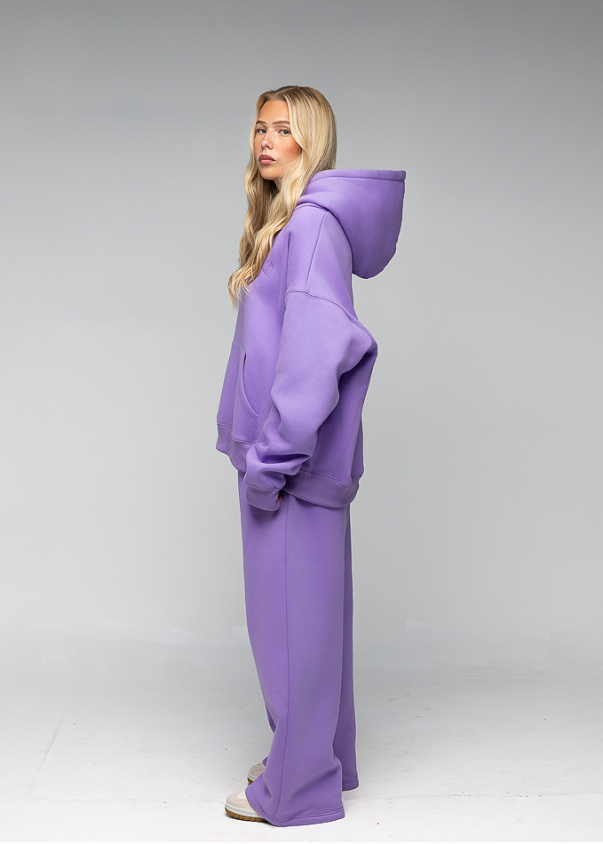 A woman modeling a matching lavender-purple lounge set consisting of an oversized zip-up hoodie and wide-leg pants, shown from a side profile against a neutral gray studio background.
