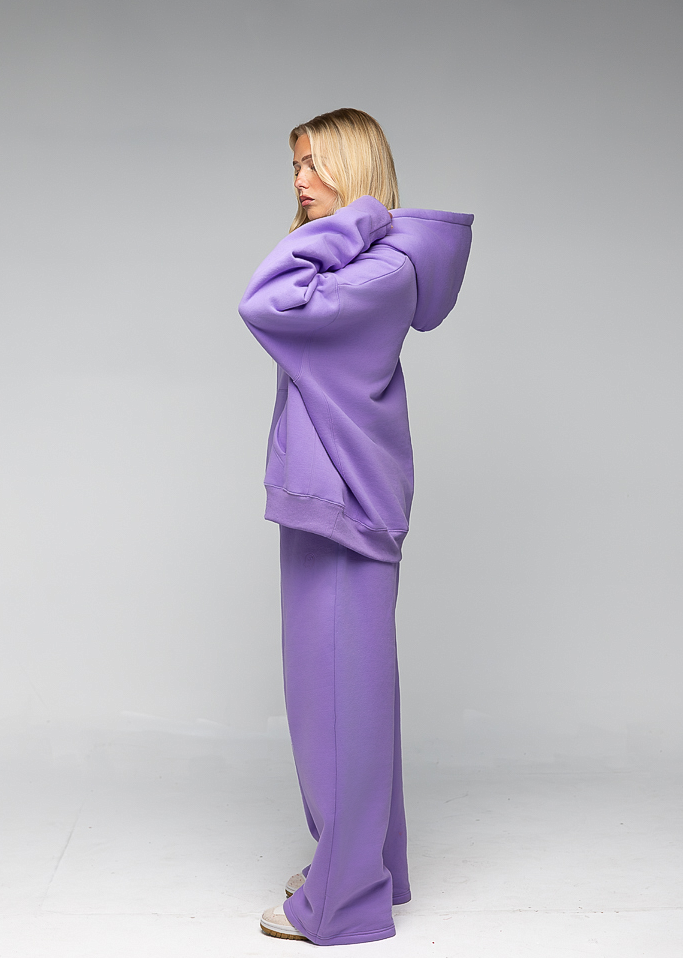 A woman modeling a matching lavender-purple lounge set consisting of a cropped hoodie and wide-leg pants, shown from a side-back angle against a neutral gray studio background.