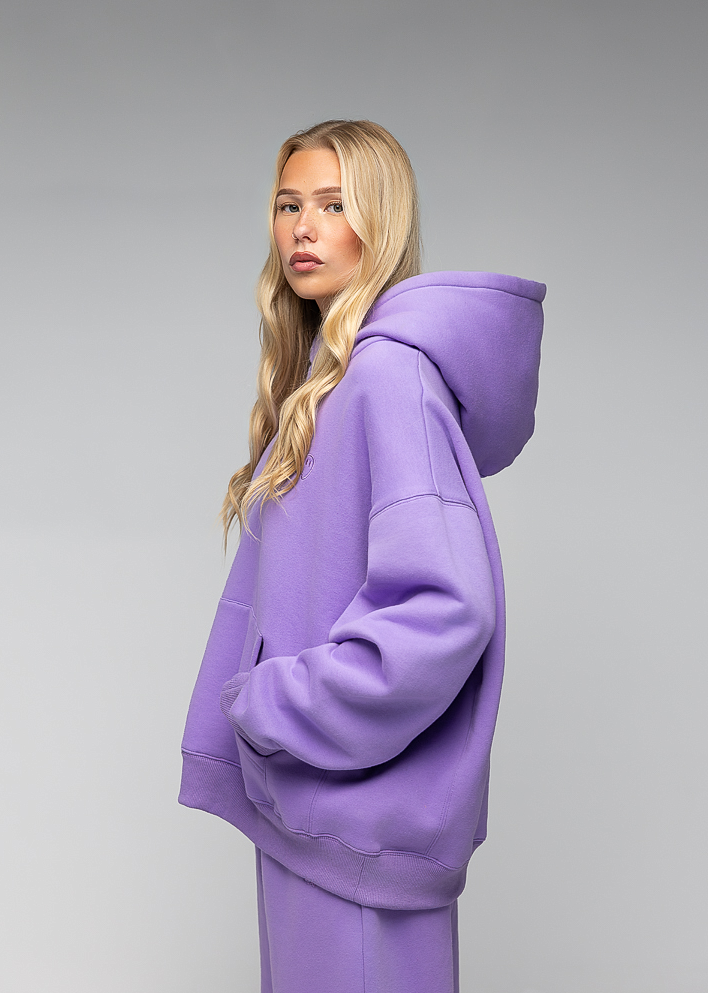 A blonde model wearing an oversized lavender-purple hoodie and matching sweatpants, shown from a side profile against a neutral gray studio background.