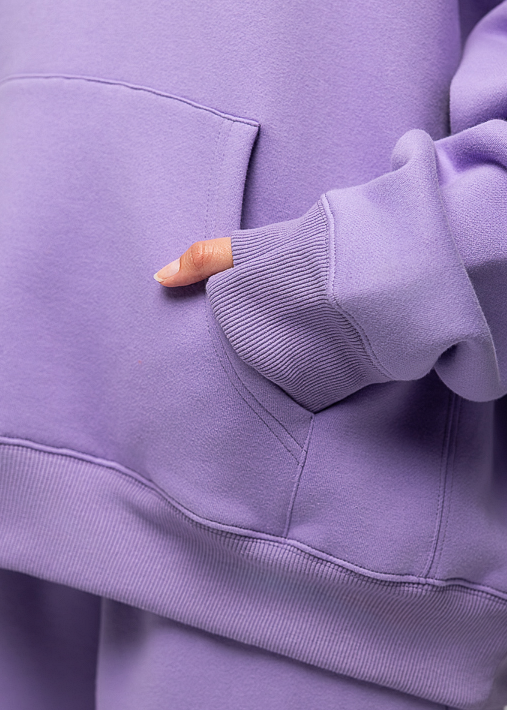 Close-up of a person wearing a lavender-purple sweatshirt and matching sweatpants, showing the sleeve, pocket, and ribbed cuffs against a plain light gray background.