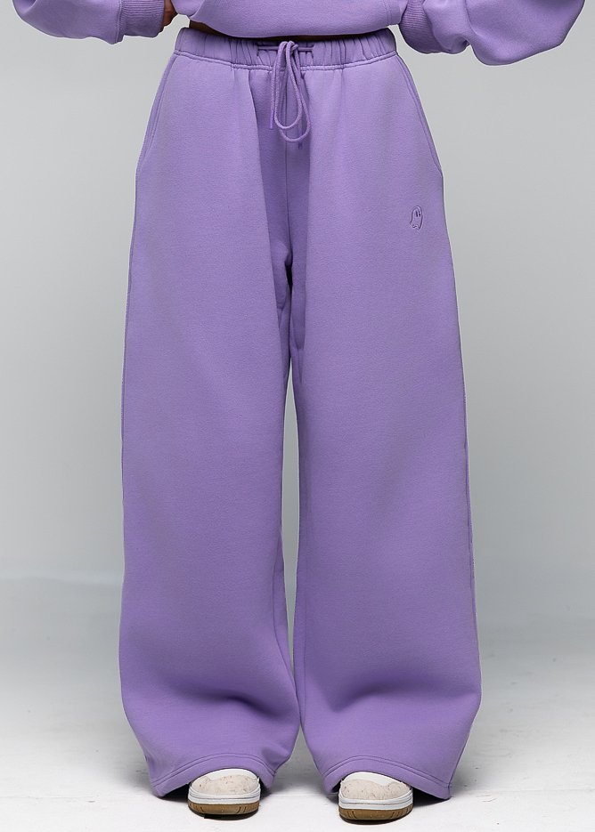A person wearing loose lavender sweatpants with a drawstring waist and a matching lavender sweatshirt, shown from the chest down against a light gray studio background.