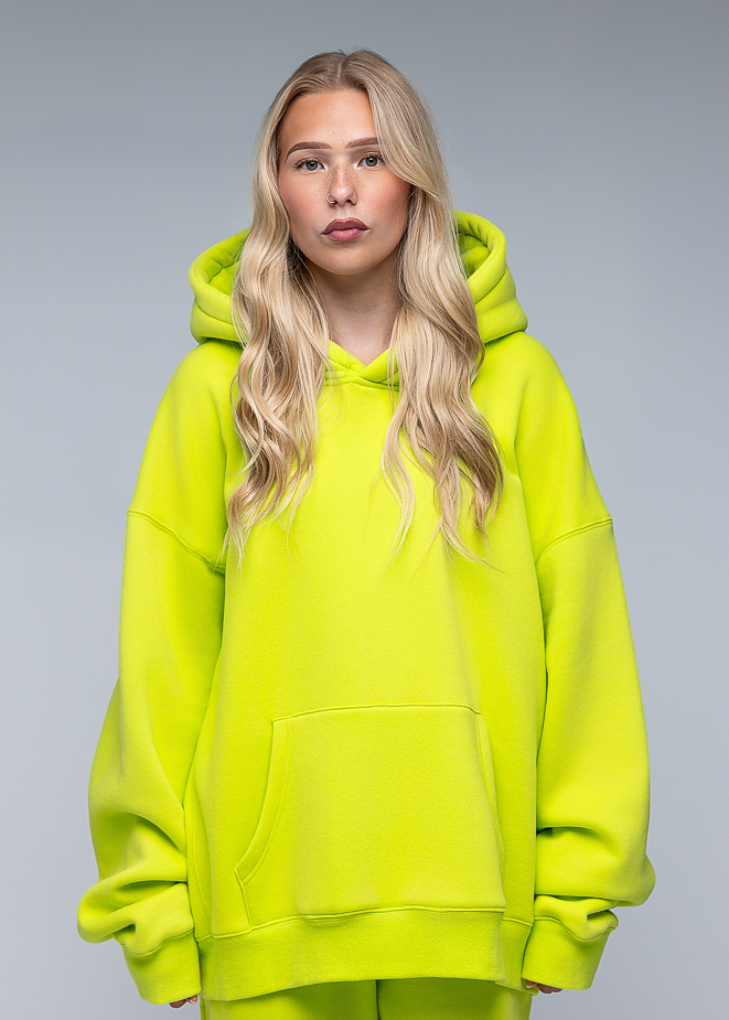 A blonde woman modeling an oversized neon yellow-green hoodie and matching sweatpants against a neutral gray studio background.