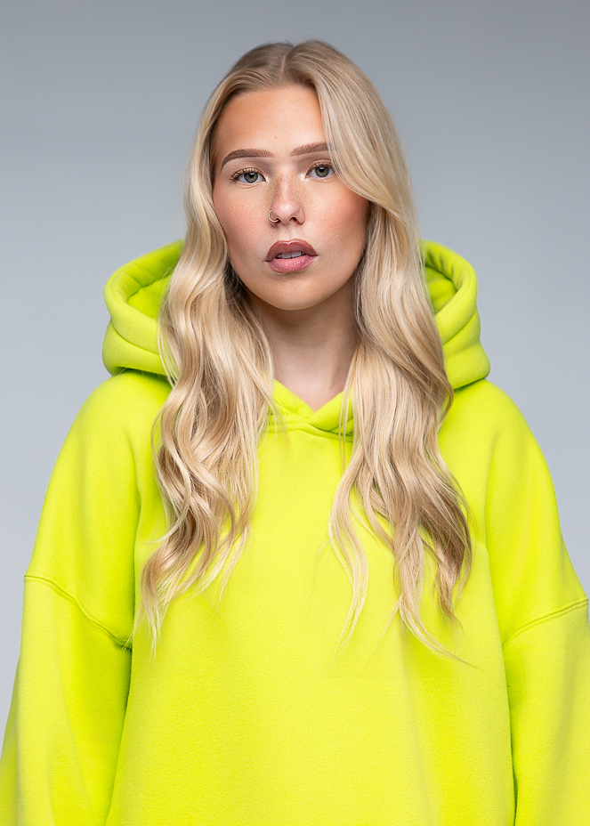 A blonde woman with long wavy hair wearing an oversized neon yellow-green hoodie, photographed against a plain light gray studio background.
