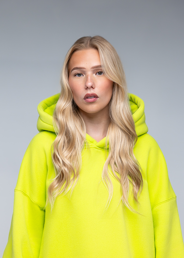 A blonde woman with long wavy hair wearing an oversized neon yellow-green hoodie, photographed against a plain light gray studio background.