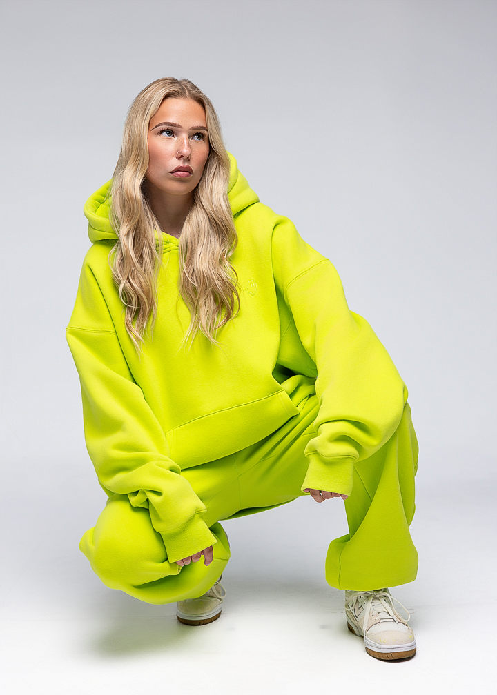 A blonde woman crouching in a neon yellow-green oversized hoodie and matching sweatpants with white sneakers, photographed against a light gray studio background.