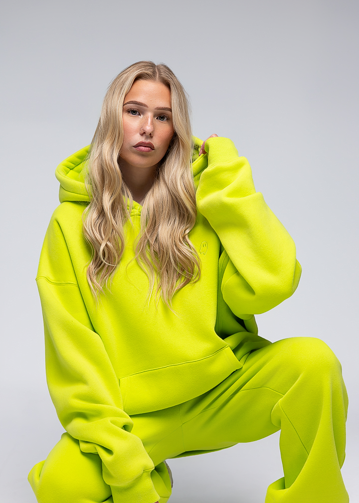 A blonde woman wearing a bright neon yellow-green matching hoodie and sweatpants set, crouching against a light gray studio background.