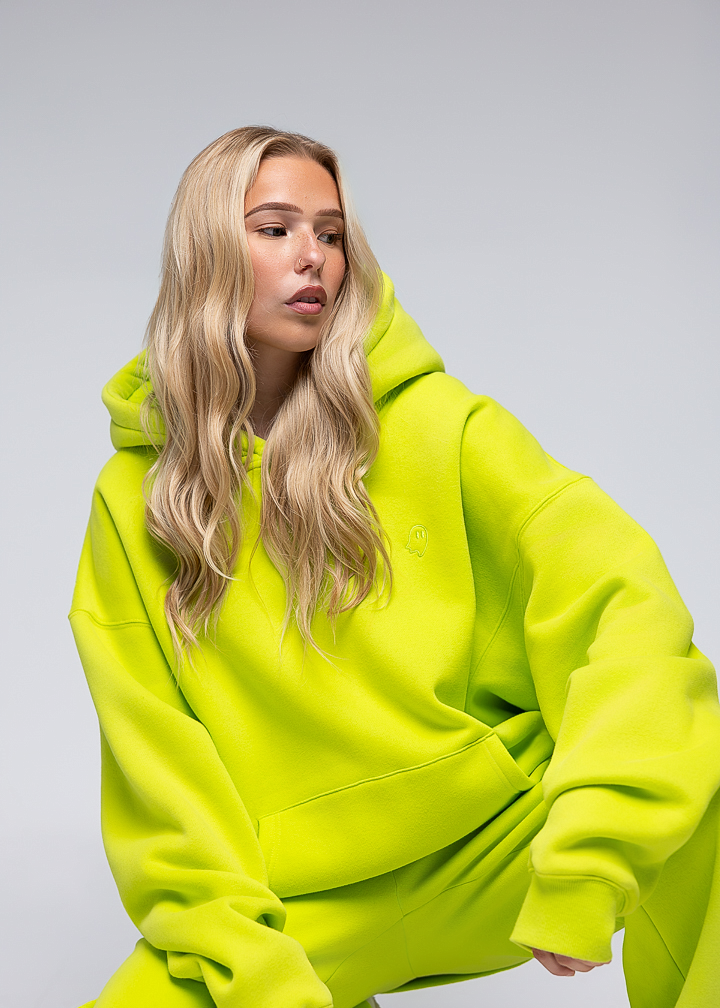 A blonde woman wearing an oversized neon yellow-green hooded sweatshirt and matching sweatpants, posed against a light gray studio background.