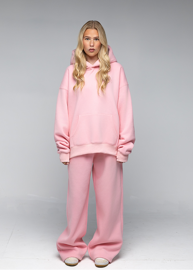 A blonde model wearing a matching light pink loungewear set consisting of an oversized pullover hoodie and wide-leg sweatpants, standing against a neutral gray studio background.
