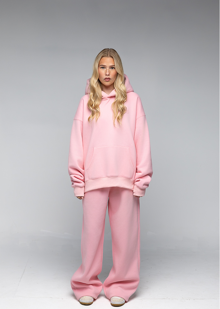 A blonde model wearing a matching light pink loungewear set consisting of an oversized pullover hoodie and wide-leg sweatpants, standing against a neutral gray studio background.
