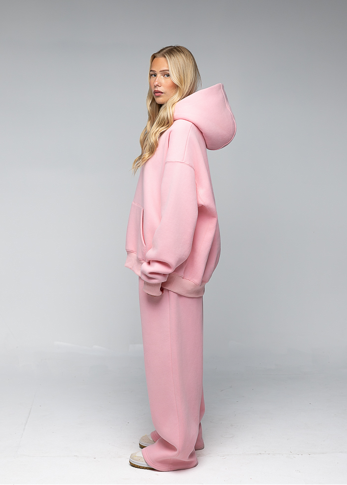 A blonde woman stands in profile wearing a matching light pink lounge set consisting of an oversized hooded sweatshirt and wide-leg sweatpants, photographed against a neutral light gray studio background.