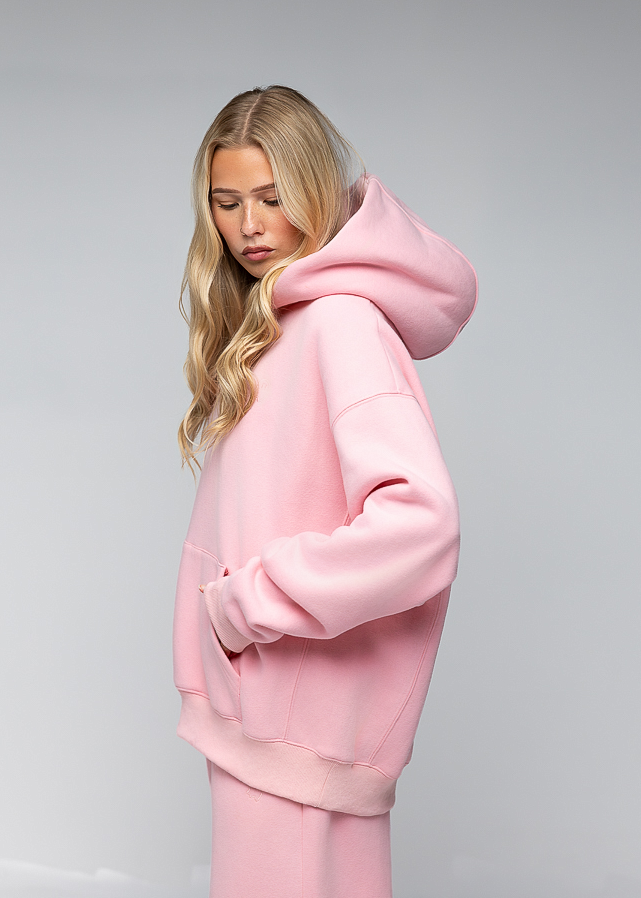 A blonde woman wearing an oversized light pink hoodie and matching pink pants, photographed against a neutral light gray studio background.