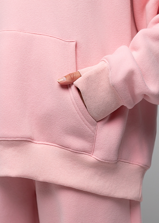 A close-up of a person wearing a soft pink sweatshirt and matching sweatpants, with the hand tucked into the front pocket of the hoodie.