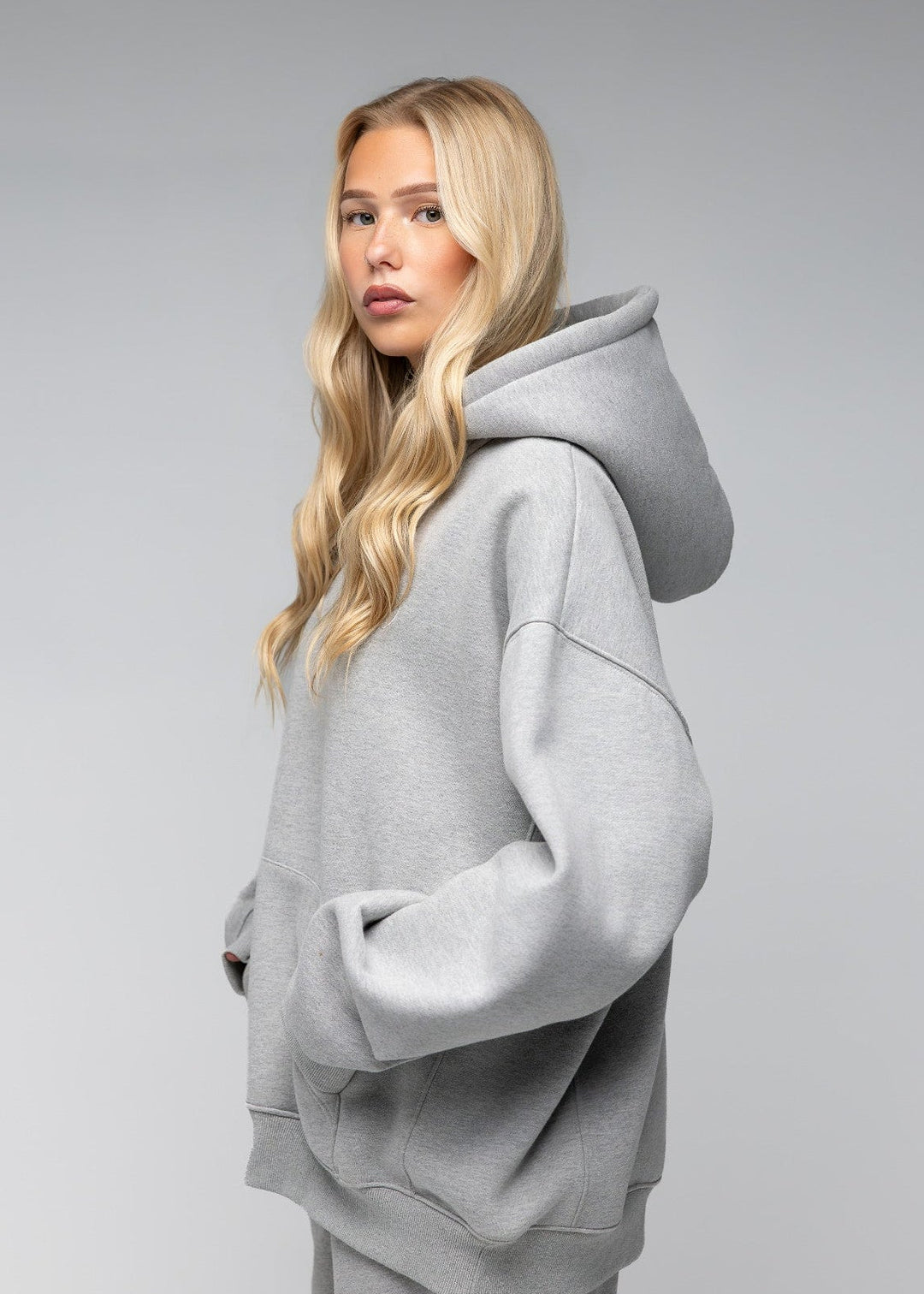 A blonde woman wearing an oversized light gray heathered hoodie and matching sweatpants, photographed against a seamless light gray studio background.