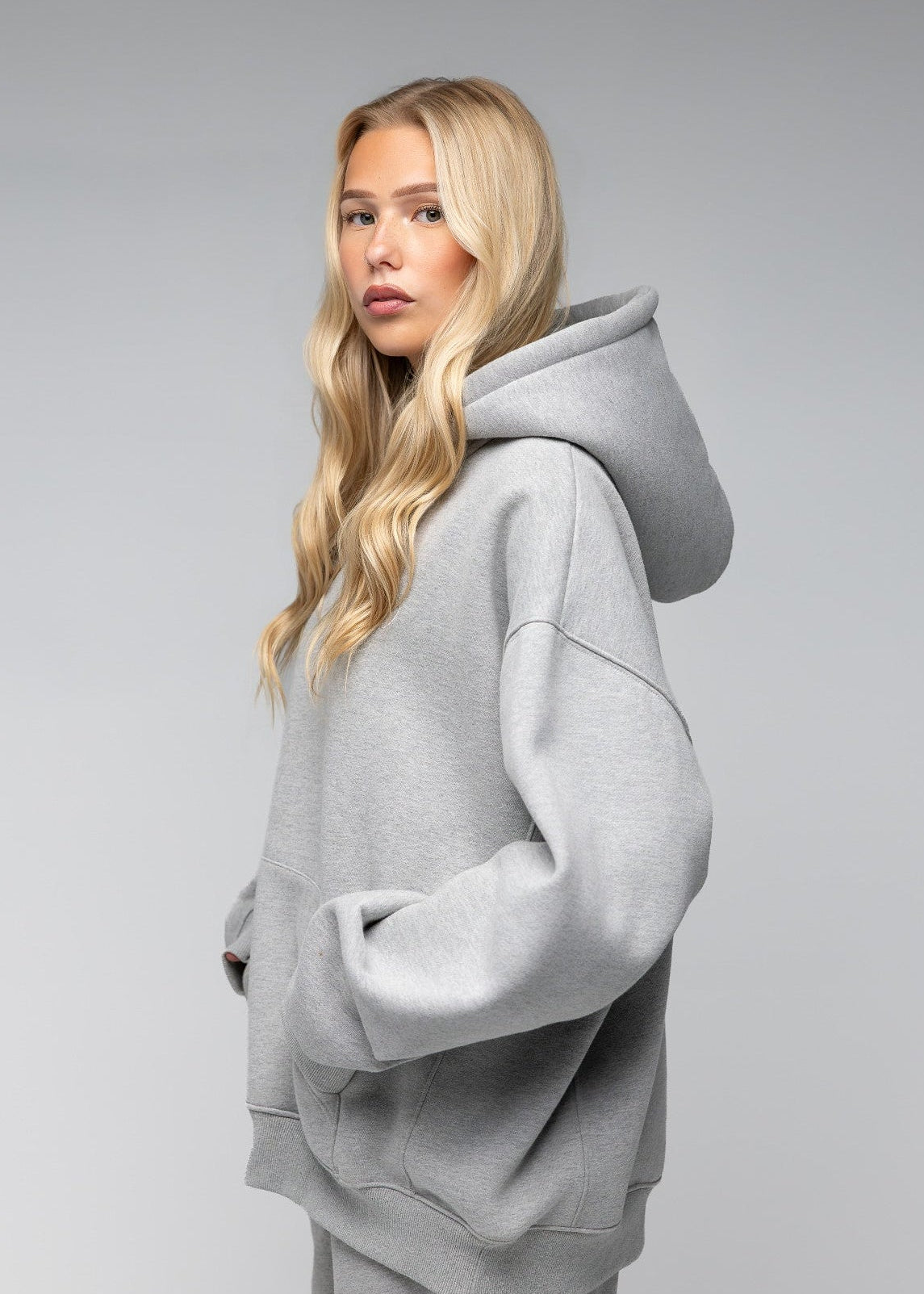 A blonde woman wearing an oversized light gray heathered hoodie and matching sweatpants, photographed against a seamless light gray studio background.