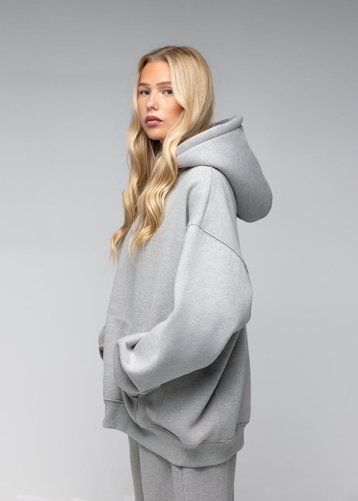 A blonde woman wearing an oversized light gray hoodie and matching sweatpants, photographed in profile against a neutral gray studio background.