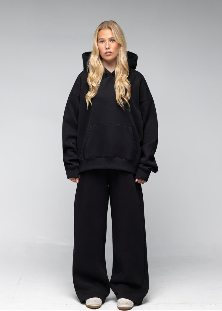 A blonde woman stands against a light gray studio background wearing an oversized black hoodie and matching black wide-leg sweatpants with white sneakers.
