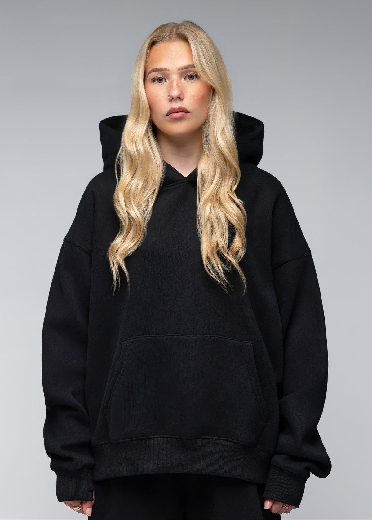 A blonde woman wearing an oversized black pullover hoodie with a front kangaroo pocket, photographed against a neutral gray studio background.