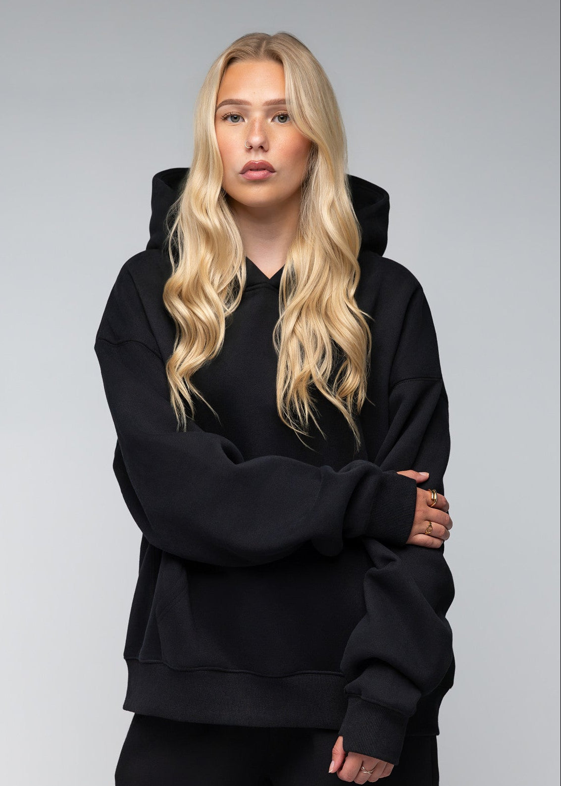 A blonde woman wearing an oversized black hoodie and matching black sweatpants, standing against a neutral gray studio background.