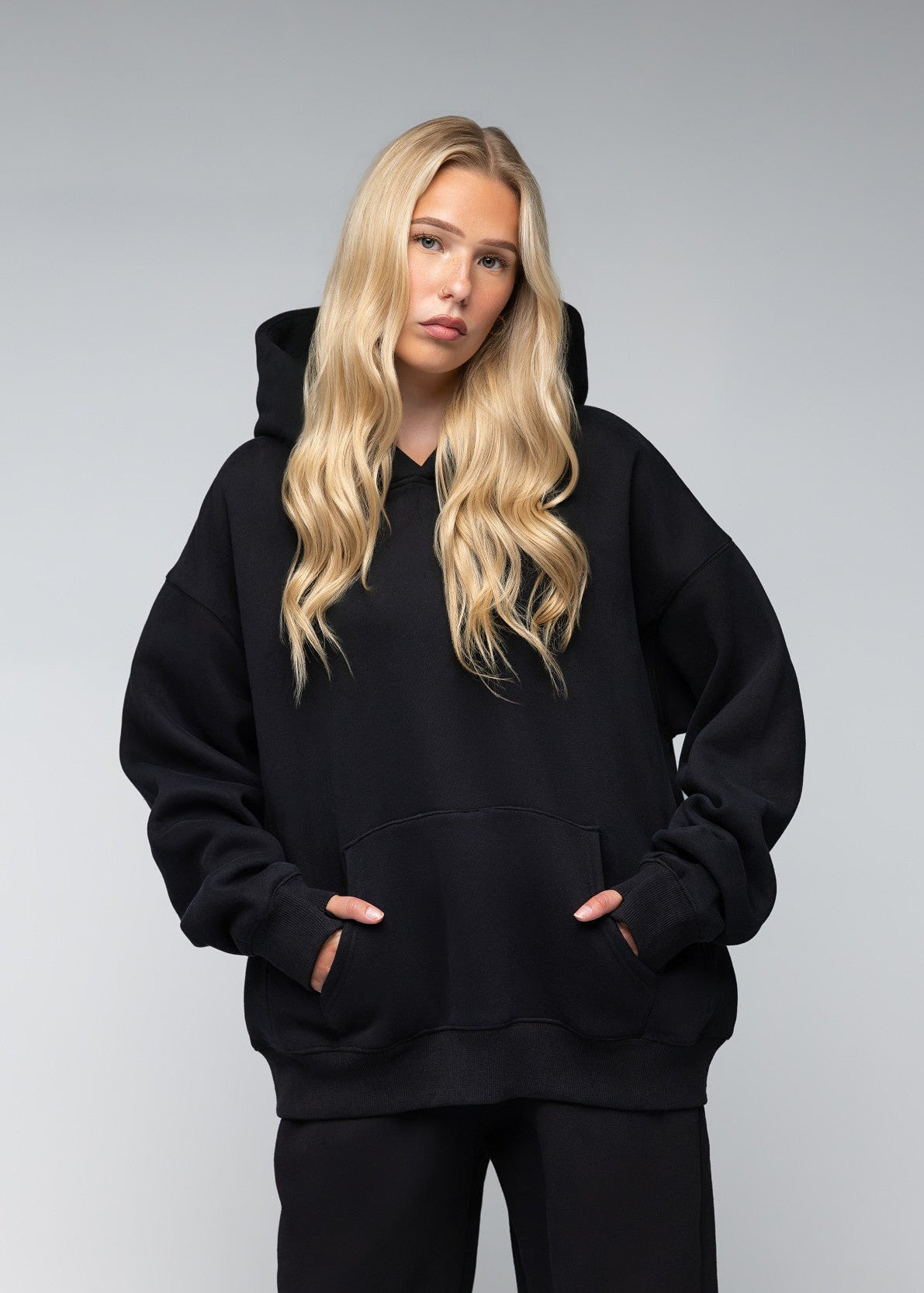 A blonde woman wearing an oversized black pullover hoodie with a front kangaroo pocket and matching black sweatpants, standing against a neutral gray studio background.