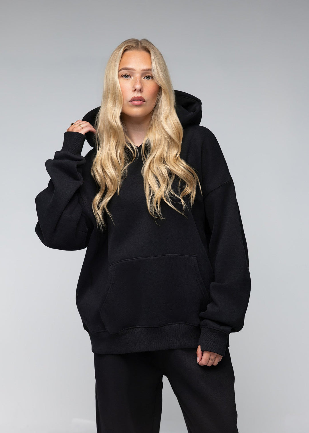 A blonde woman modeling an oversized black hoodie with matching black sweatpants against a light gray studio background.