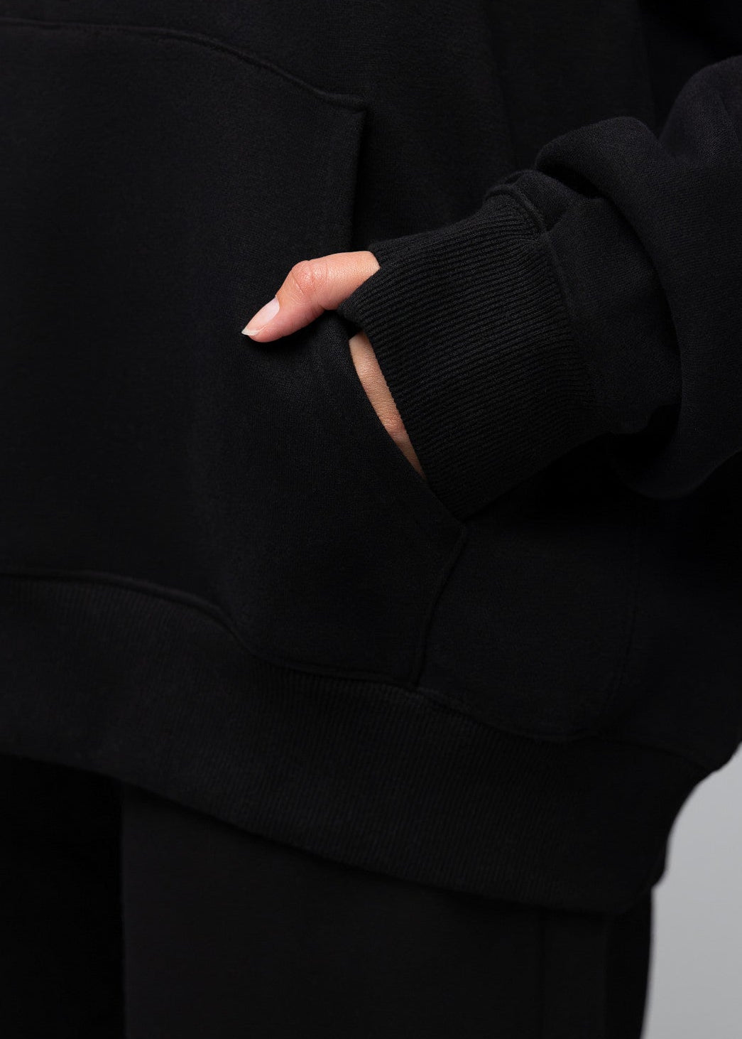 Close-up of a person wearing a black hoodie with a front kangaroo pocket, shown from the chest down with one hand in the pocket, against a light gray background.