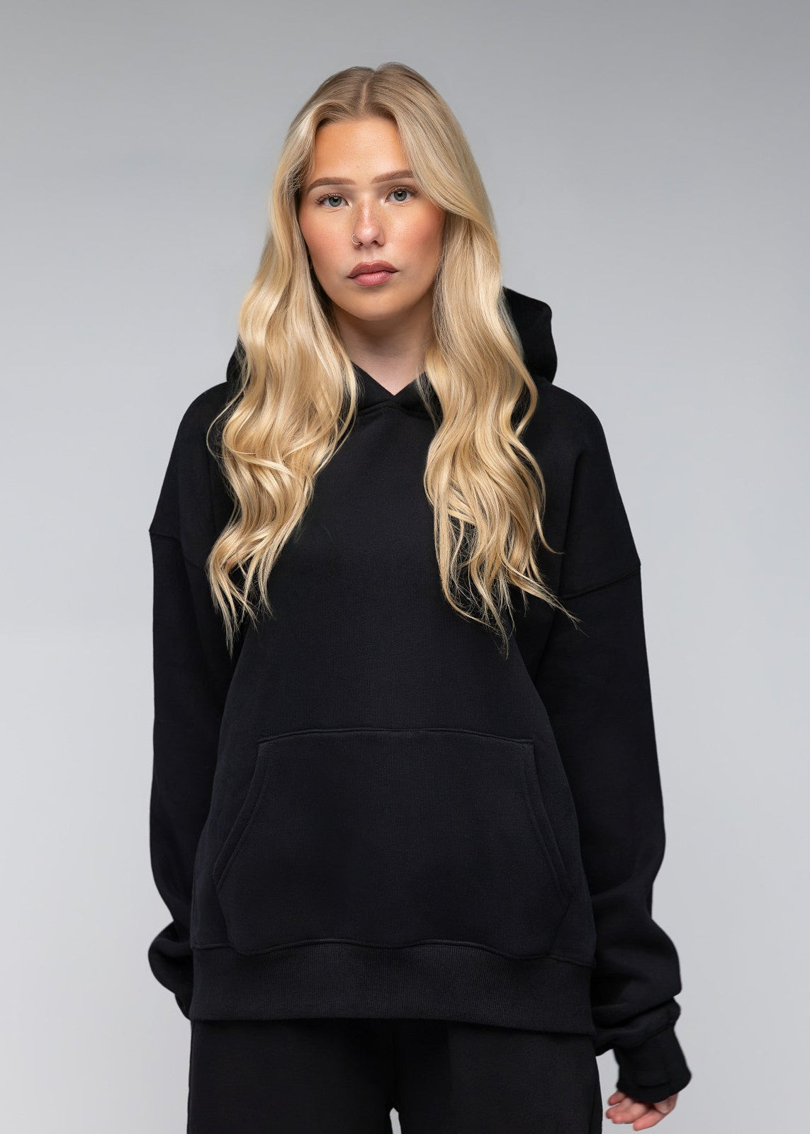 A blonde woman modeling a black pullover hoodie with a front kangaroo pocket, paired with black sweatpants, against a neutral gray studio background.