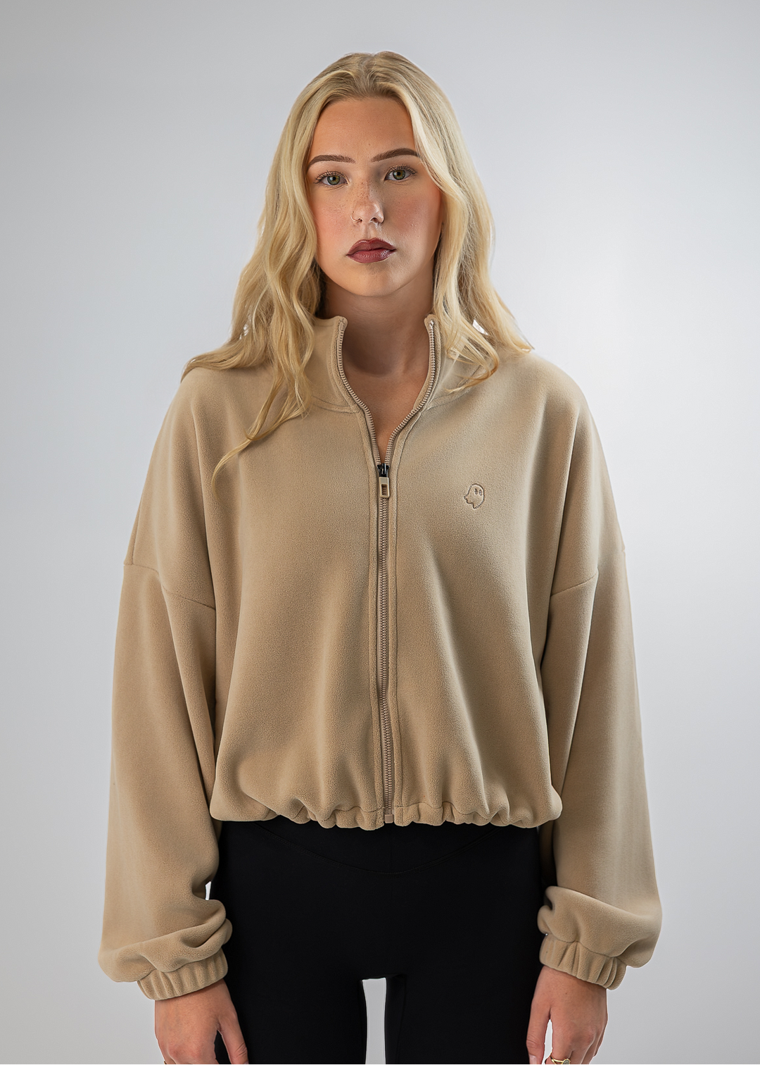 A blonde woman wearing a beige cropped zip-up fleece jacket with elastic cuffs and a small embroidered logo on the chest, paired with black pants, against a light gray studio background.