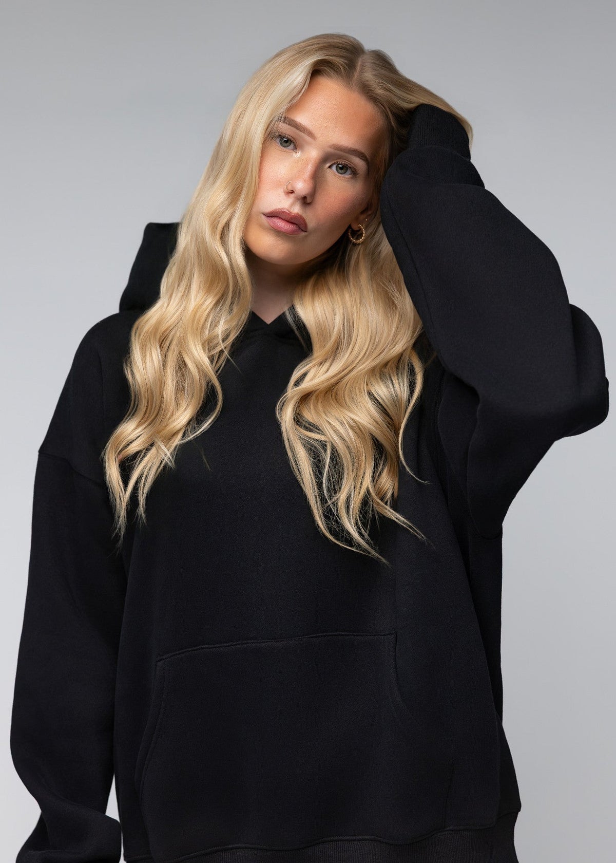 A blonde woman wearing an oversized black pullover hoodie, posing against a neutral gray studio background.