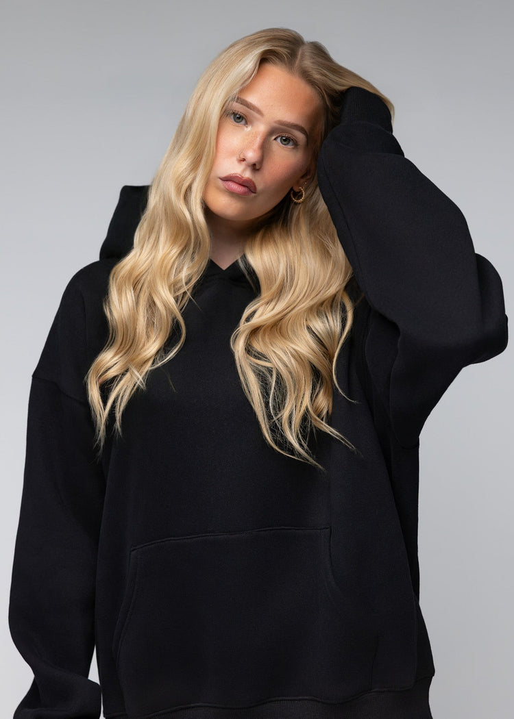 A blonde woman wearing an oversized black pullover hoodie, posing against a neutral gray studio background.