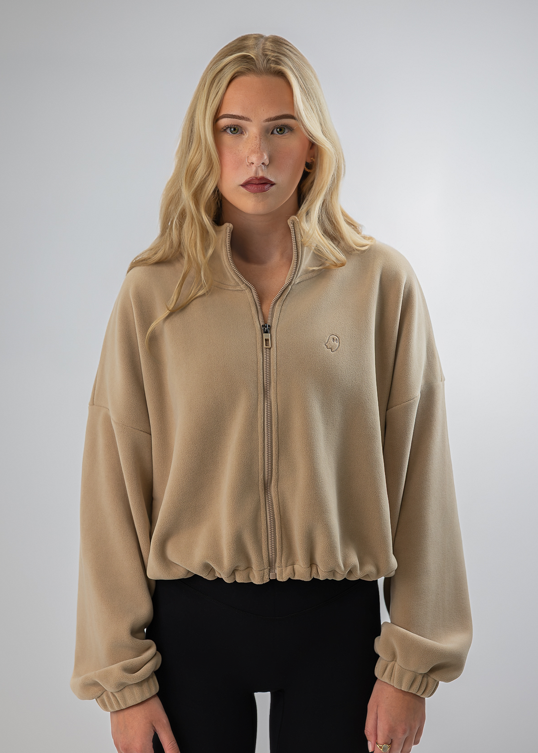A blonde model wearing a beige cropped zip-up fleece jacket with elastic cuffs and a small embroidered logo on the chest, paired with black pants, against a neutral gray studio background.