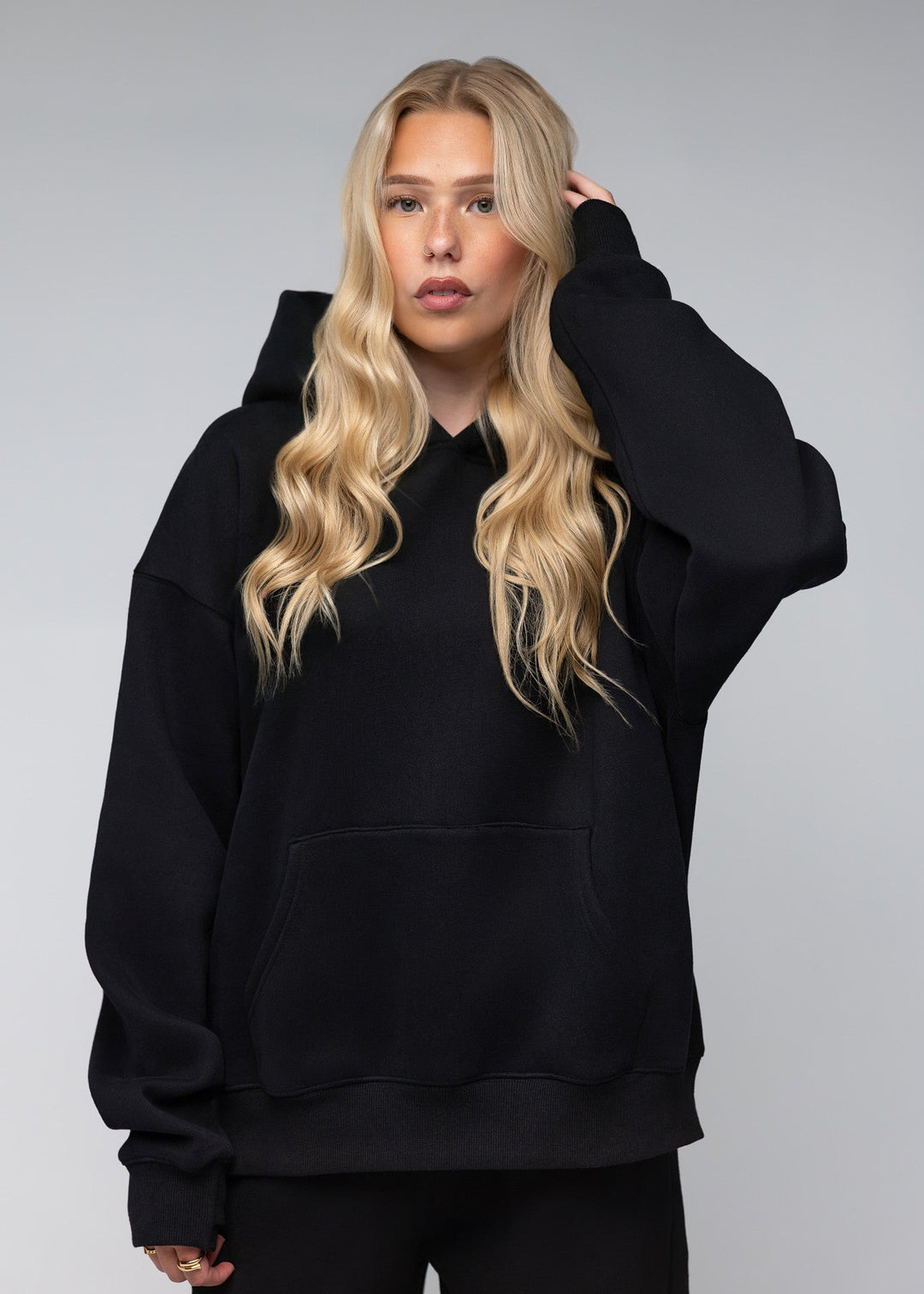 A blonde woman wearing an oversized black pullover hoodie with a front kangaroo pocket, photographed against a neutral gray studio background.