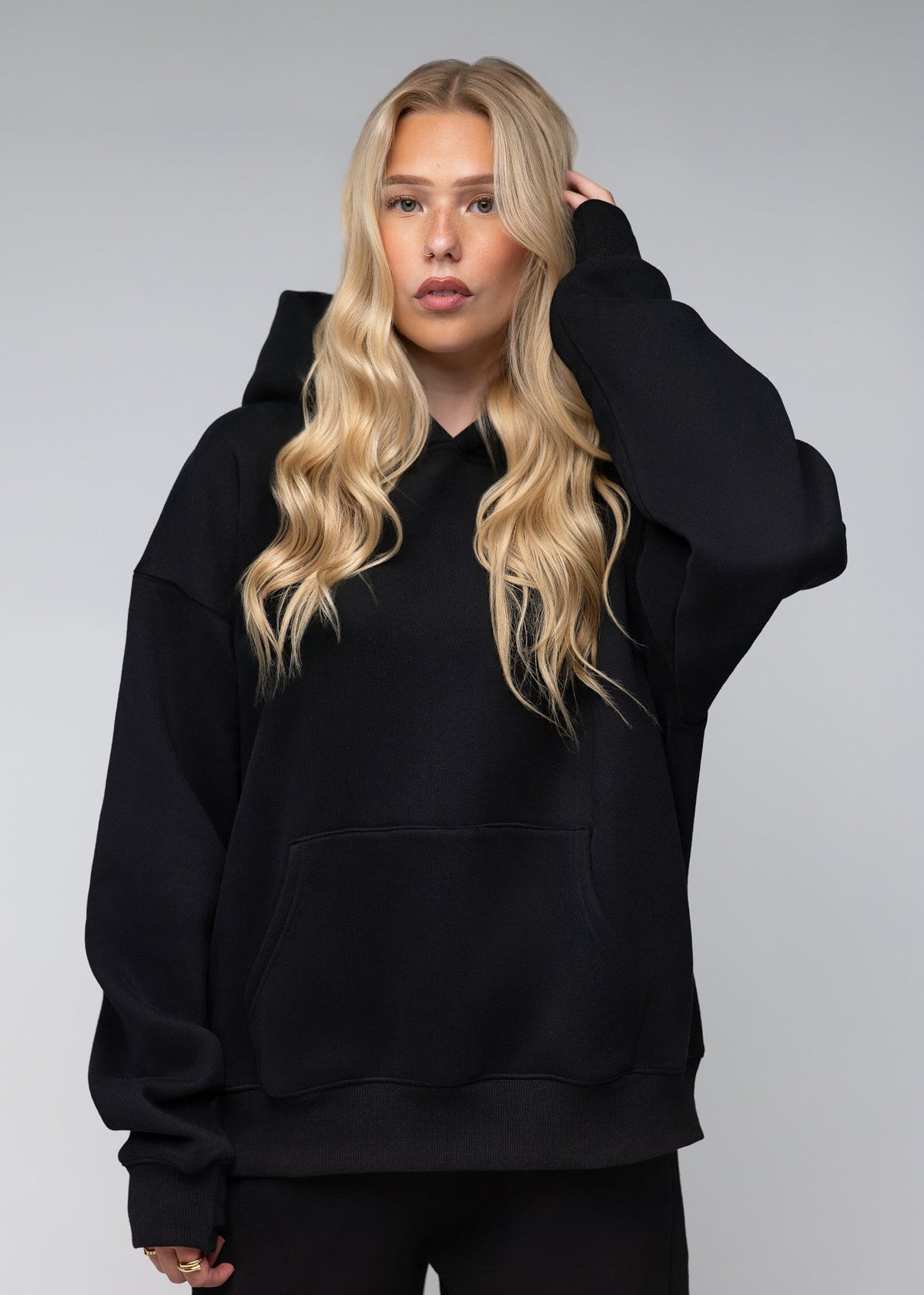 A blonde woman wearing an oversized black pullover hoodie with a front kangaroo pocket, photographed against a neutral gray studio background.