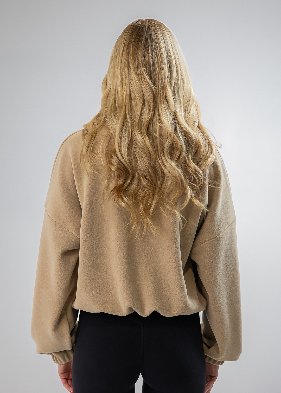 A woman viewed from behind wearing a beige cropped sweatshirt with voluminous wavy blonde hair and black pants, set against a plain light gray studio background.