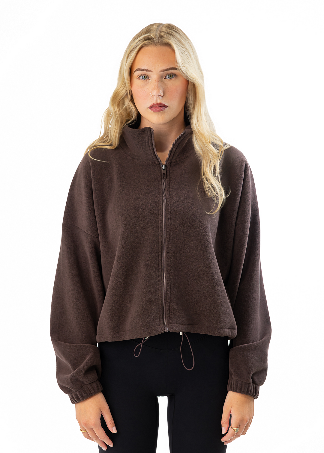 A blonde woman wearing a dark brown cropped zip-up fleece jacket with a stand collar and drawstring hem, paired with black pants, photographed against a solid white background.