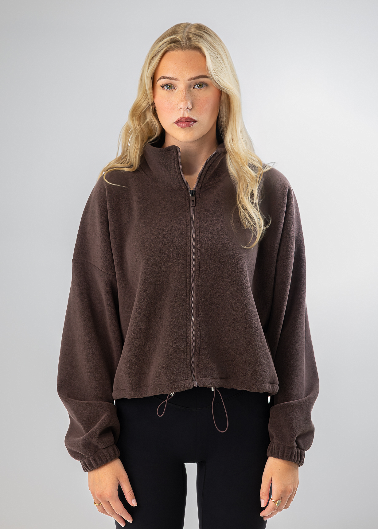A blonde woman modeling a dark brown cropped zip-up fleece jacket with a stand collar and drawstring hem, paired with black pants, against a light gray studio background.