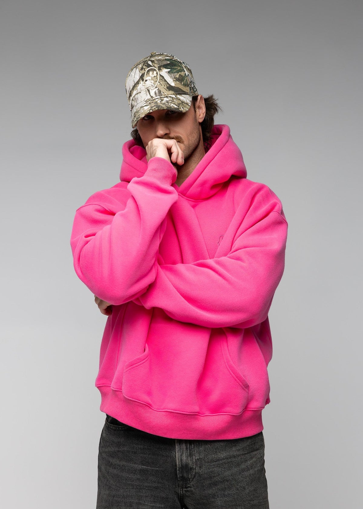 A man wearing a bright hot pink oversized hoodie, dark gray jeans, and a camouflage baseball cap, posing with his arms crossed in front of a solid gray studio background.