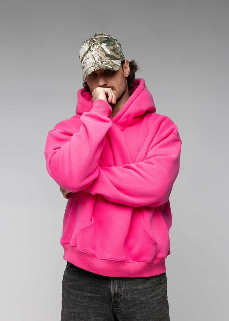 A man wearing a bright hot pink oversized hoodie, dark gray jeans, and a camouflage baseball cap, posing with his arms crossed in front of a solid gray studio background.