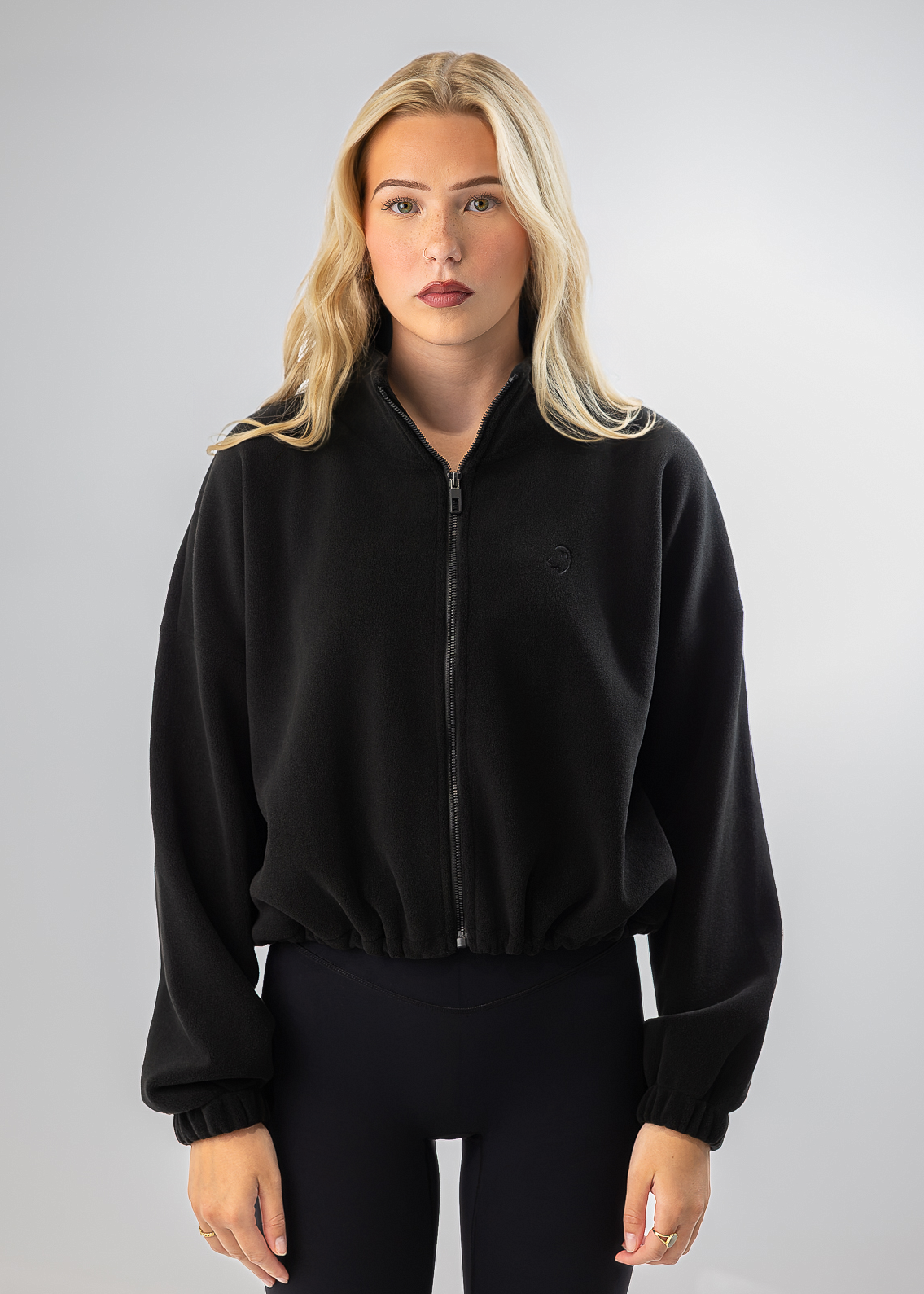 A blonde woman modeling a black zip-up fleece jacket with a small tonal embroidered logo on the left chest, paired with black leggings, against a light gray studio background.