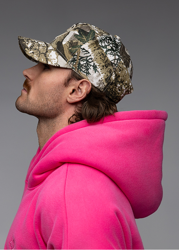 A man in profile wearing a bright pink hoodie and a camouflage baseball cap, photographed against a smooth gray studio background.