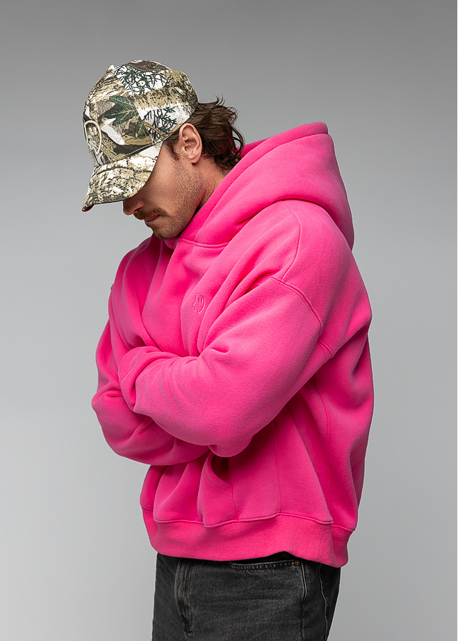 A man wearing a bright pink oversized hoodie, dark gray jeans, and a camouflage baseball cap, standing against a neutral gray studio background.